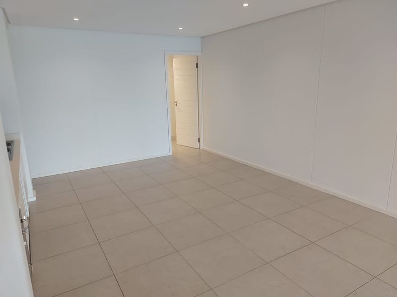 A bright, empty open-plan living area featuring neutral tiled flooring, recessed downlighting, and a clean, modern finish throughout.