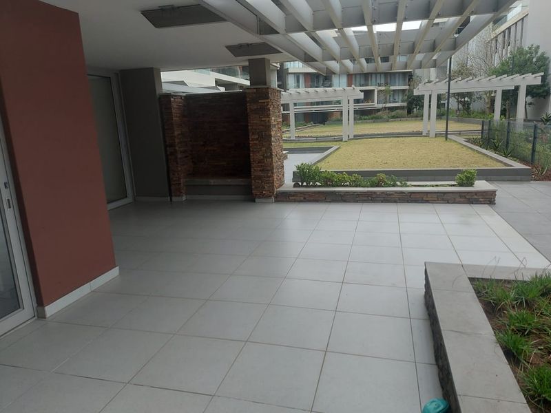 Spacious covered patio featuring tiled flooring, a modern pergola, and stone-clad built-in seating, overlooking a landscaped communal garden area.