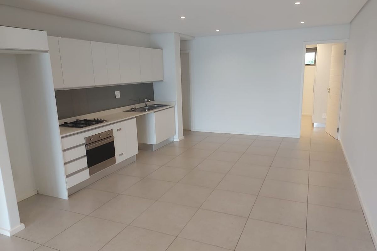 Modern open-plan kitchen and living area featuring sleek white cabinetry, integrated appliances, neutral tiled flooring, and recessed lighting in a clean, vacant space.
