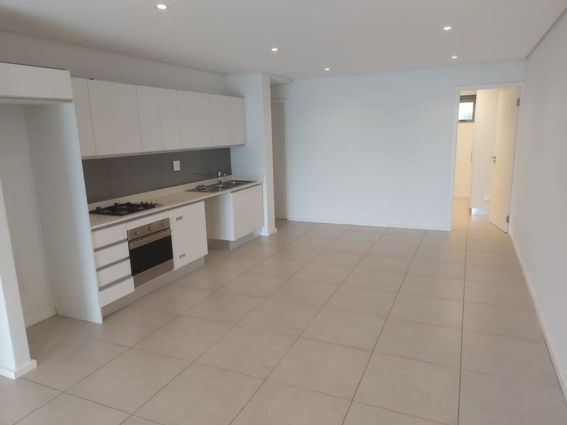 Modern open-plan kitchen and living area featuring sleek white cabinetry, integrated appliances, neutral tiled flooring, and recessed lighting in a clean, vacant space.