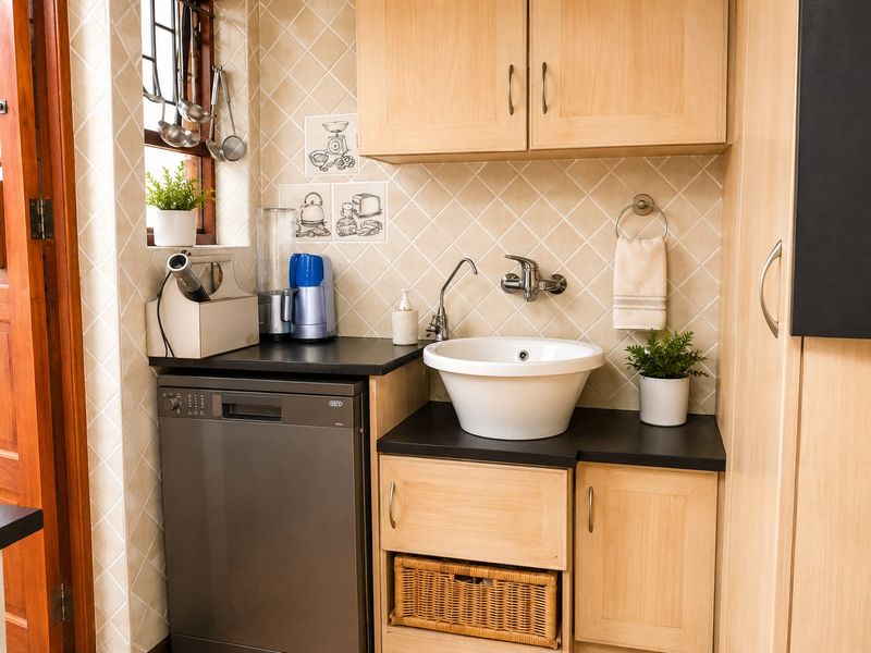 A functional scullery area featuring light wood cabinetry, a dark countertop, a vessel sink, and a built-in dishwasher with tiled backsplash.