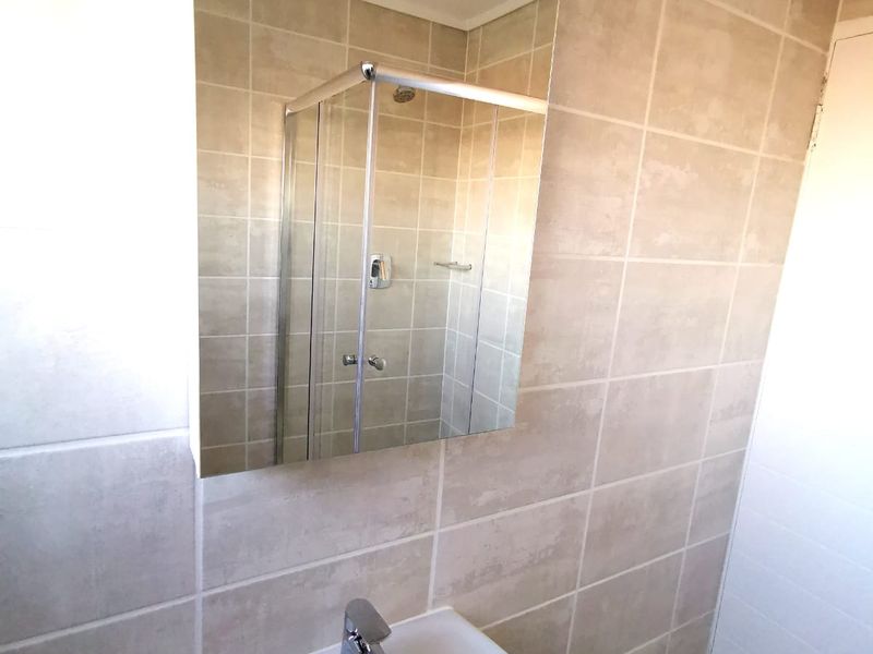 A clean, functional bathroom featuring a pedestal basin with chrome mixer tap, wall-mounted mirror, and a glass-enclosed shower with neutral ceramic tiling.