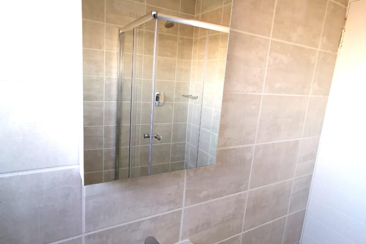 A clean, functional bathroom featuring a pedestal basin with chrome mixer tap, wall-mounted mirror, and a glass-enclosed shower with neutral ceramic tiling.