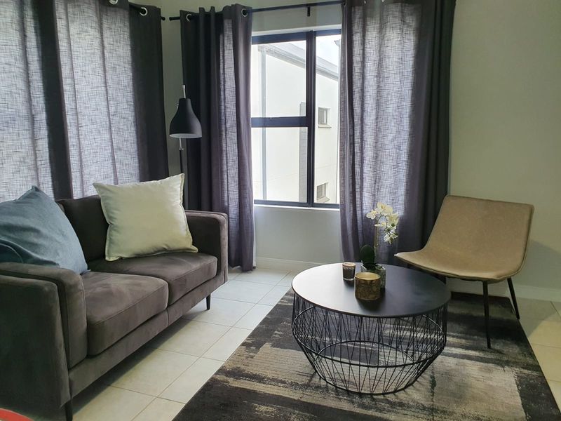 A bright living room featuring tiled flooring and a large window with dark curtains, providing natural light and a view of the adjacent building.