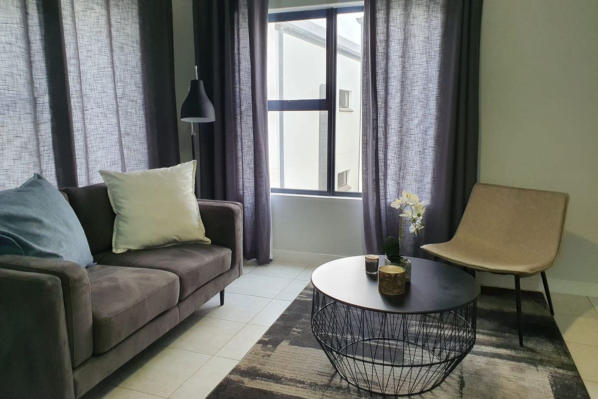 A bright living room featuring tiled flooring and a large window with dark curtains, providing natural light and a view of the adjacent building.