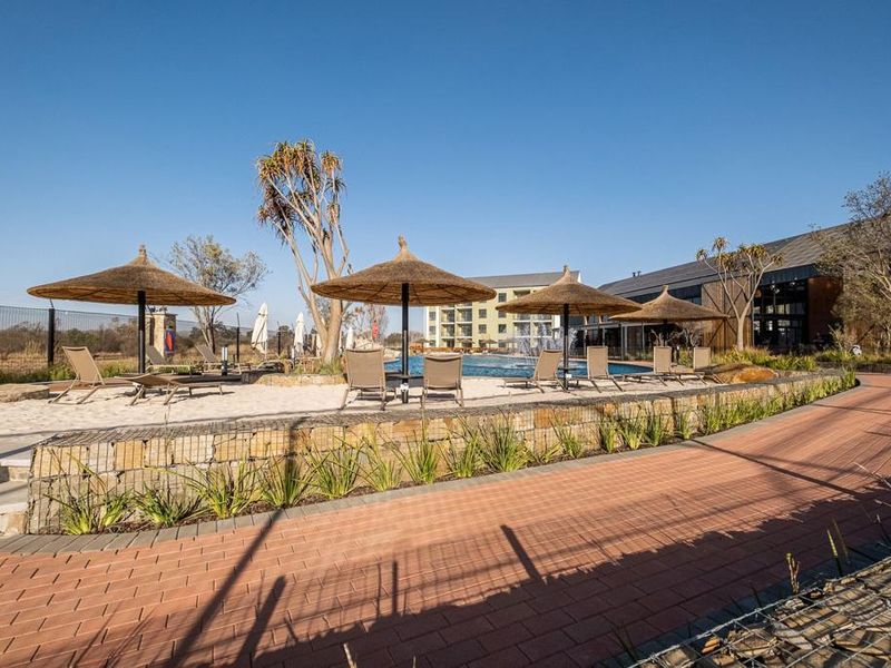 A well-maintained communal pool area featuring a stone retaining wall, paved walkways, and a sandy lounging section with thatched umbrellas.