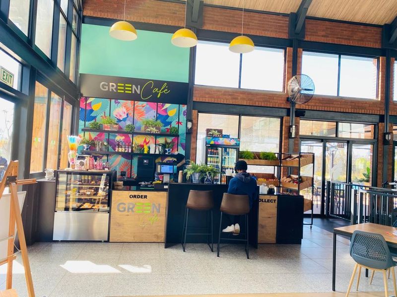 A modern, high-ceilinged cafe space featuring exposed brick walls, large windows for natural light, tiled flooring, and a functional service counter area.