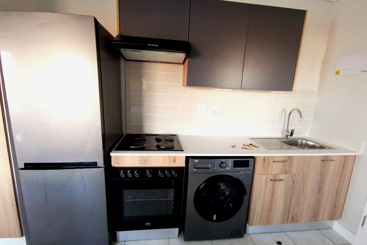 A compact, modern kitchen featuring wood-finish cabinetry, a stainless steel sink, built-in oven with electric hob, extractor fan, and tiled backsplash.