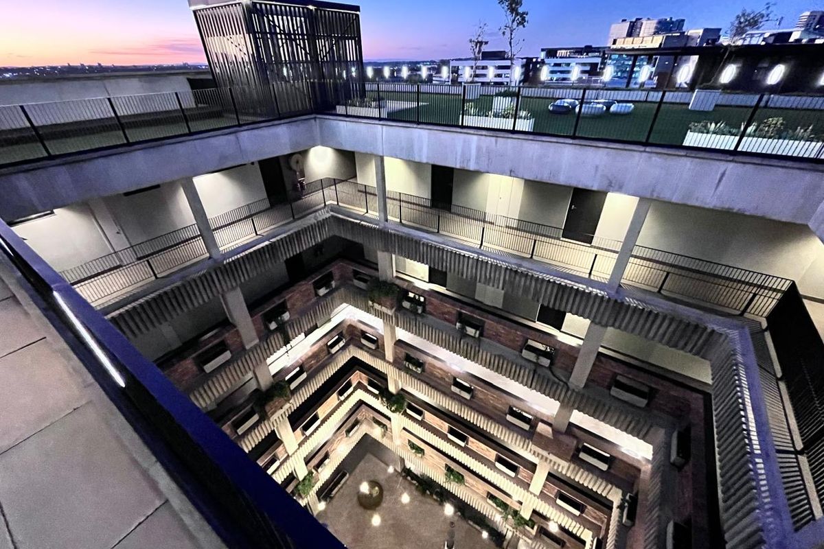 An elevated aerial view of a modern apartment complex featuring a central atrium, open-air walkways, and a rooftop terrace at dusk.