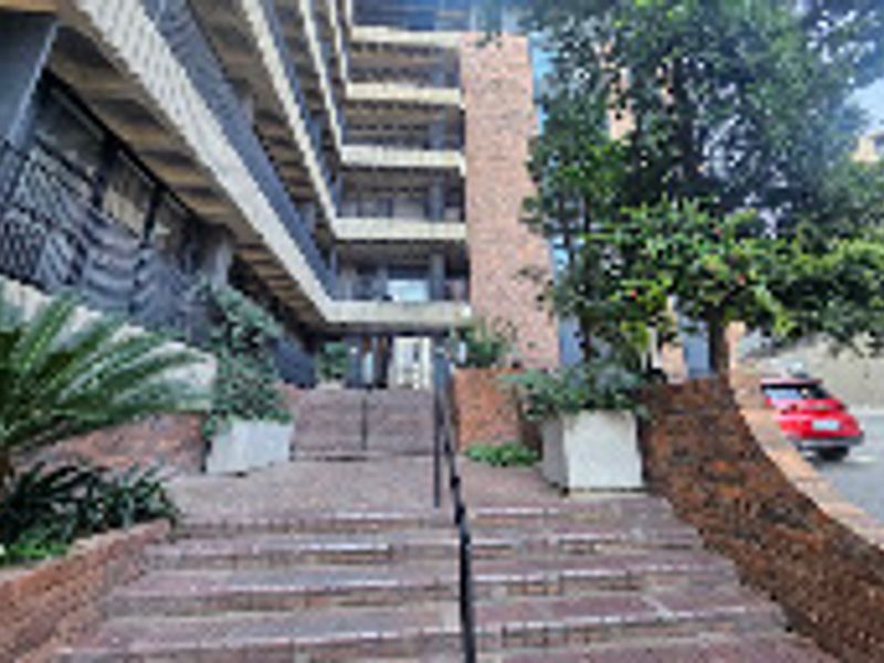 Exterior view of the apartment building entrance featuring a brick staircase, metal handrails, and established concrete planters leading to the main building facade.