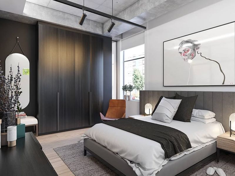 Modern bedroom featuring a sleek built-in wardrobe, exposed concrete ceiling with track lighting, and large windows providing ample natural light.