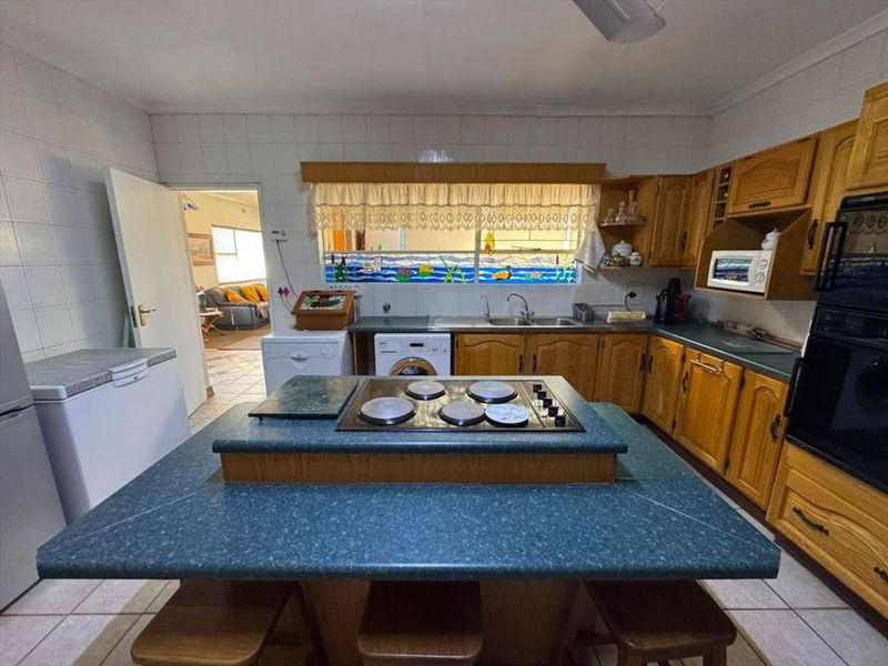 A functional kitchen featuring wooden cabinetry, a central island with an integrated electric hob, built-in oven, and tiled finishes throughout.