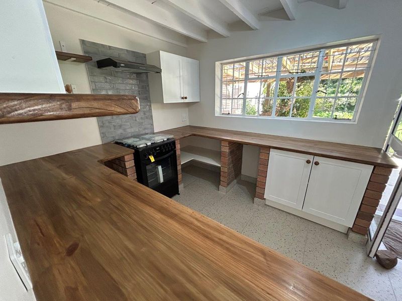 A bright kitchen featuring wooden countertops, exposed brick supports, built-in oven, extractor fan, and exposed ceiling beams with a view to the garden.