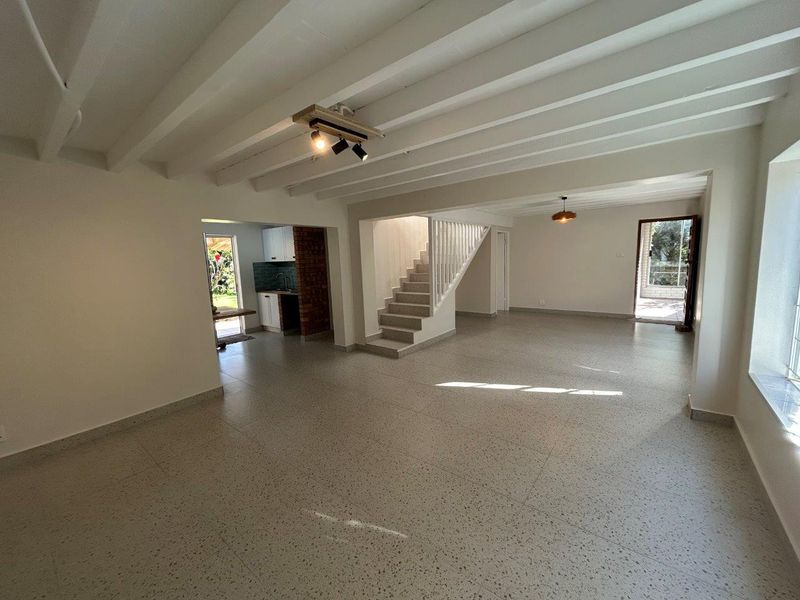 Spacious open-plan living area featuring exposed beam ceilings, terrazzo flooring, and a central staircase, with an adjacent kitchen area visible.