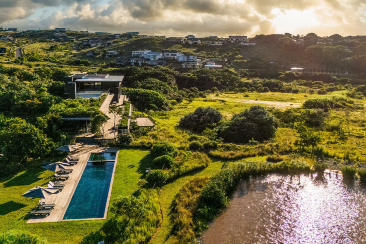 Aerial view of a modern property featuring a long swimming pool, timber decking, and expansive landscaped gardens overlooking a natural pond and surrounding estate.