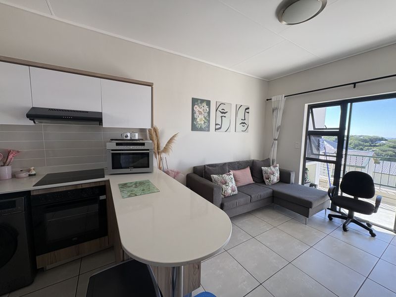 Modern open-plan apartment living area featuring a compact kitchen with built-in appliances, tiled flooring, and direct access to a balcony through a sliding door.