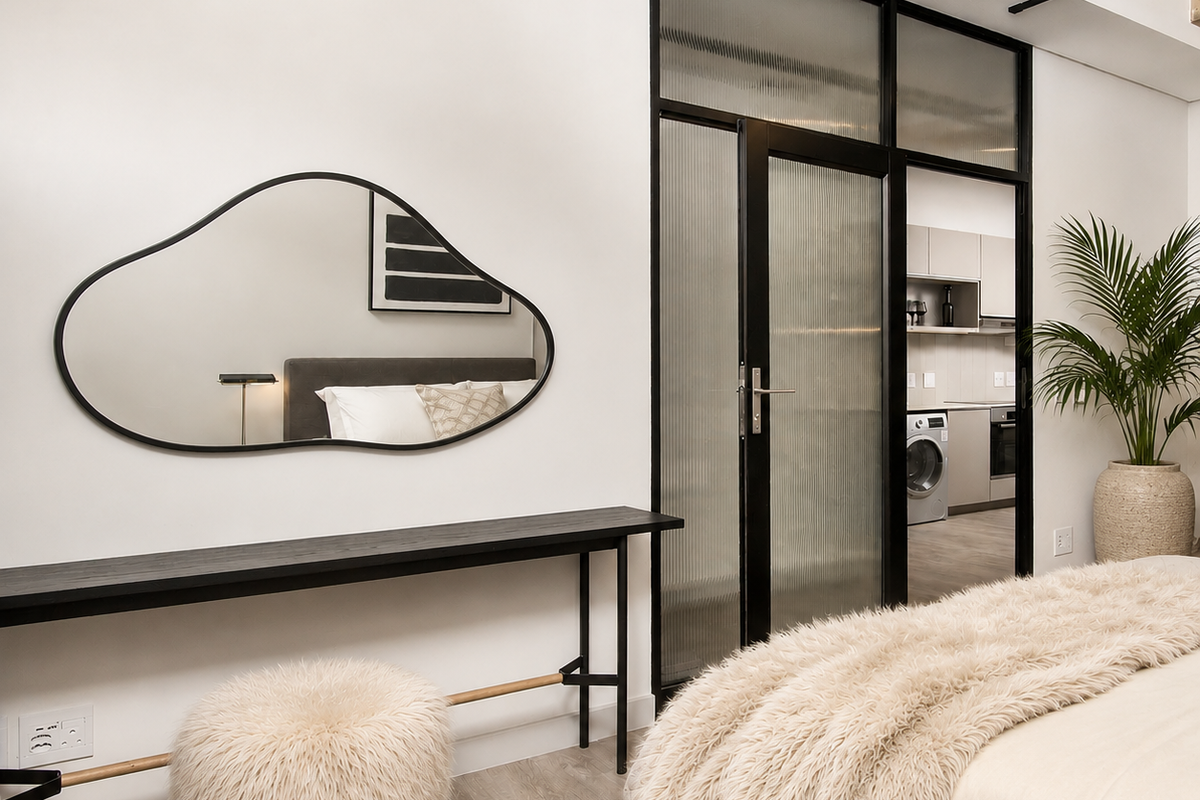 Modern bedroom featuring laminate flooring, an exposed concrete ceiling beam, and a stylish black metal-framed glass door leading to an adjacent kitchen area.