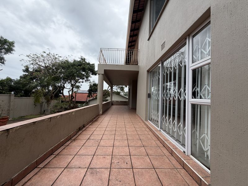 A spacious covered balcony featuring tiled flooring, sliding glass doors with security gates, and an elevated railing overlooking the garden area.