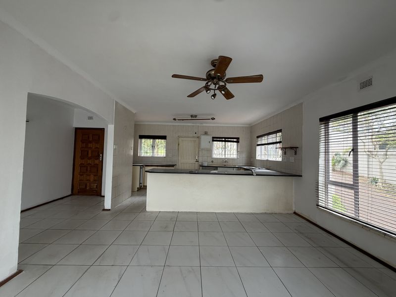 An open-plan living and kitchen area featuring tiled flooring, a central kitchen island, ceiling fan, and large windows with venetian blinds.