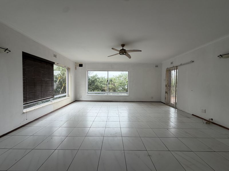 Spacious, empty living area featuring neutral tiled flooring, a ceiling fan, and large windows providing natural light and garden views.