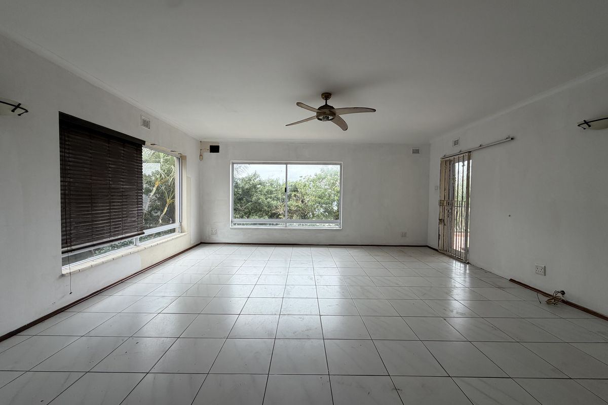 Spacious, empty living area featuring neutral tiled flooring, a ceiling fan, and large windows providing natural light and garden views.
