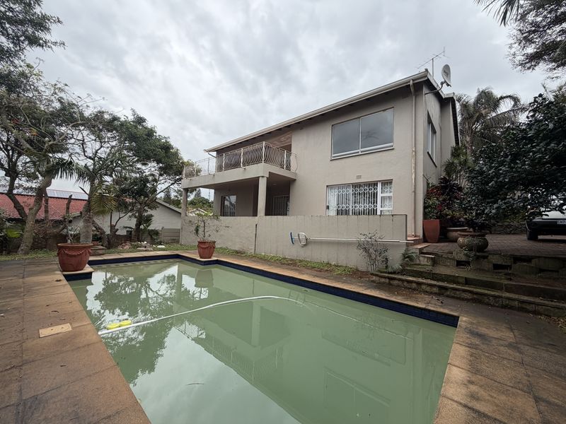 Rear exterior view of a two-story house featuring a swimming pool, paved deck area, and an upper-level balcony with wrought iron railings.