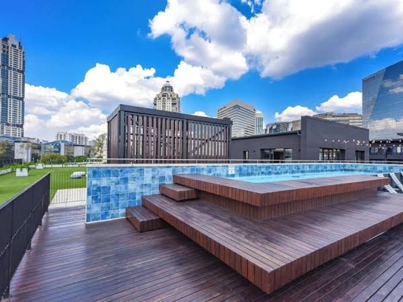 A modern rooftop pool area featuring raised timber decking, a tiled pool surround, and a timber privacy screen with expansive city skyline views.