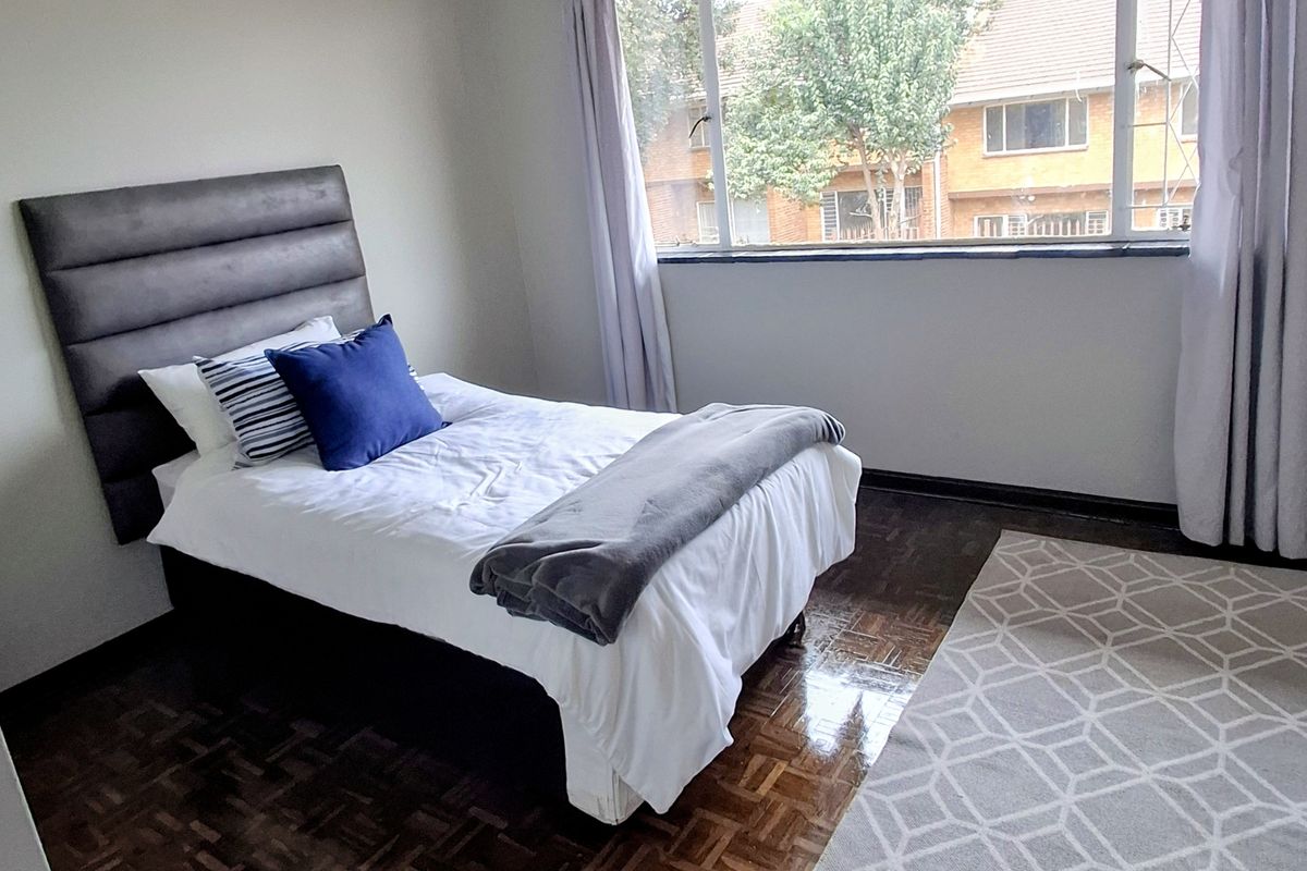 A bedroom featuring classic parquet flooring, neutral wall tones, and a large window providing natural light and a view of the surrounding complex.