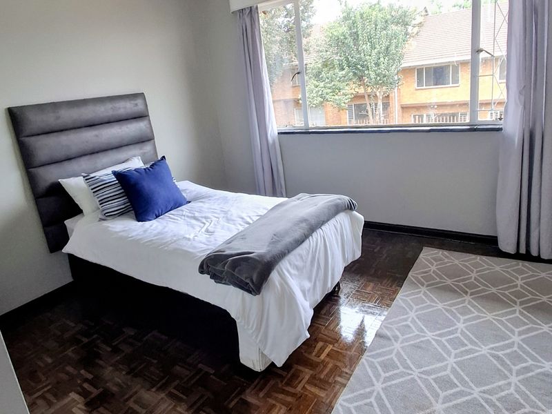 A bedroom featuring classic parquet flooring, neutral wall tones, and a large window providing natural light and a view of the surrounding complex.