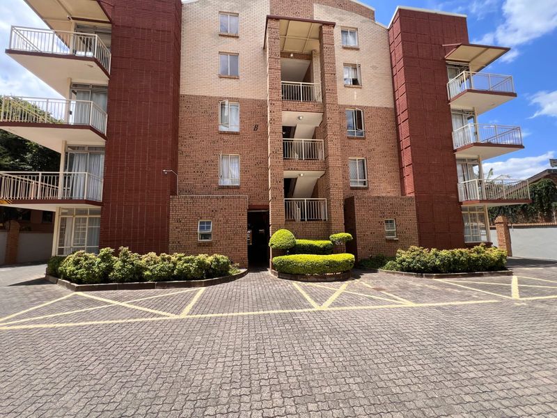 Front elevation of a multi-story apartment building featuring brick facade, private balconies, and a paved parking area with landscaped garden beds.