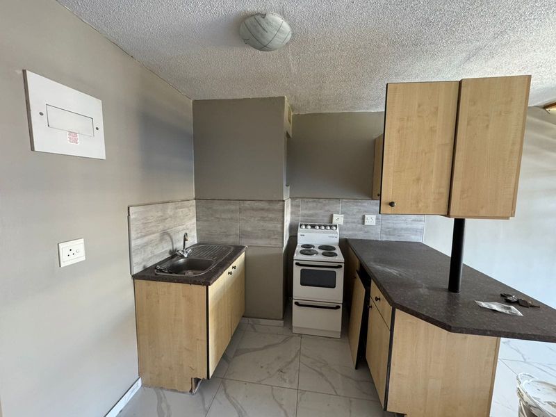 A compact kitchen featuring light wood-finish cabinetry, dark laminate countertops, a tiled backsplash, and a freestanding stove, currently in need of minor repairs.