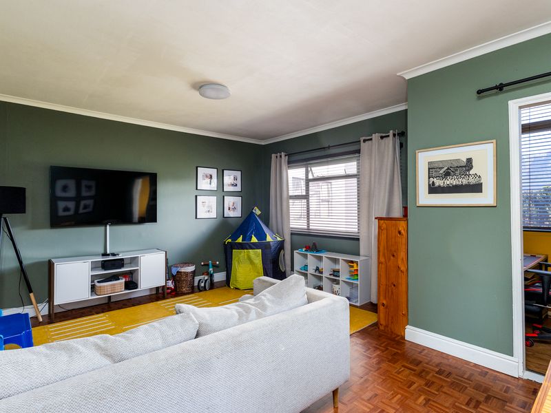 A well-maintained living room featuring classic parquet flooring, sage green accent walls, and natural light through a window with fitted blinds.