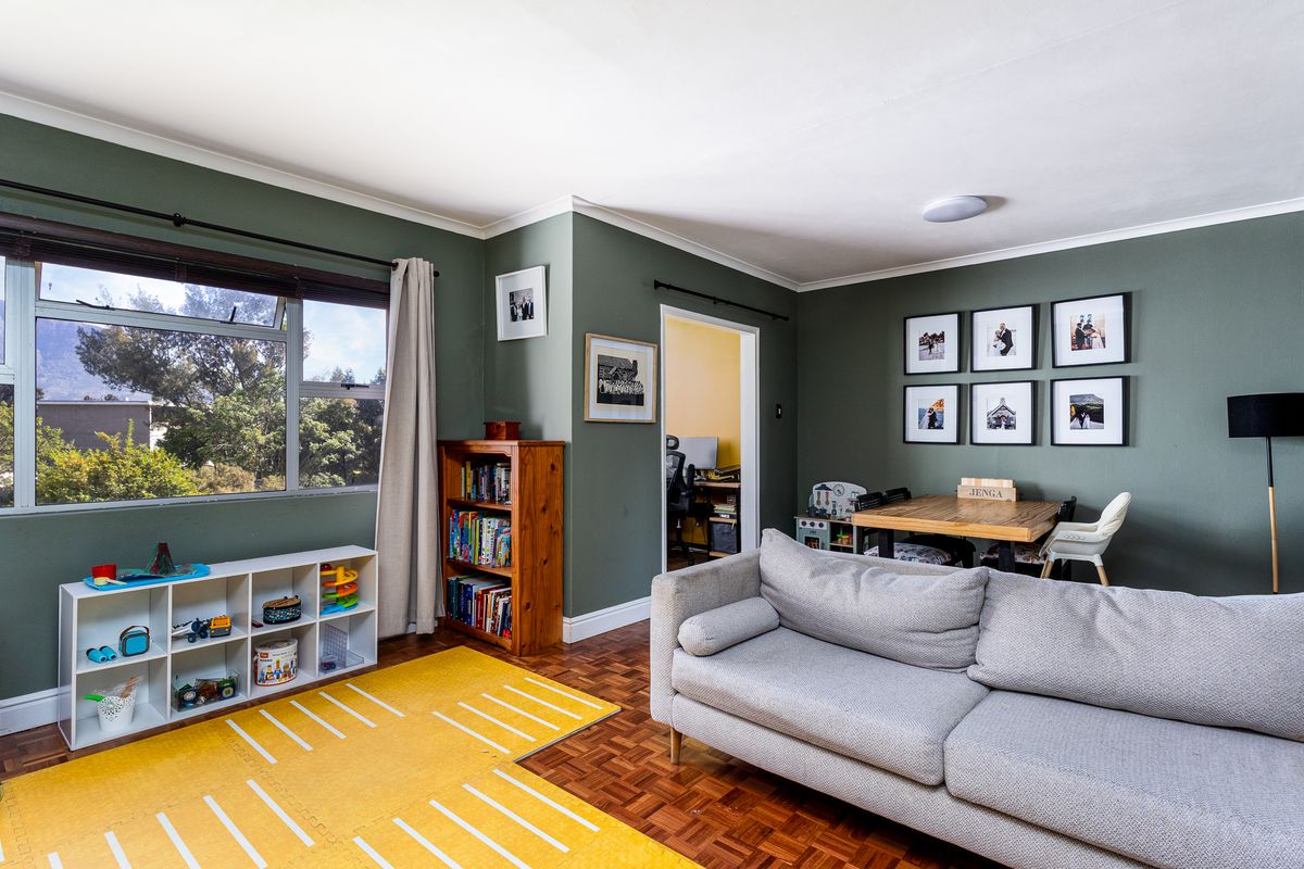 A well-maintained living room featuring classic parquet flooring, coved cornicing, and large windows providing natural light and pleasant exterior views.