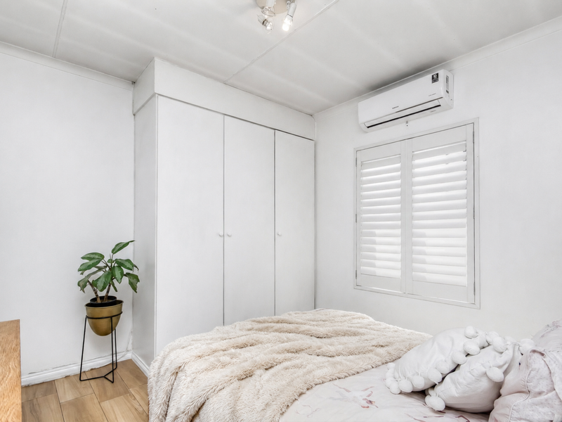 A bright bedroom featuring built-in cupboards, an air conditioning unit, plantation shutters, and modern laminate flooring.