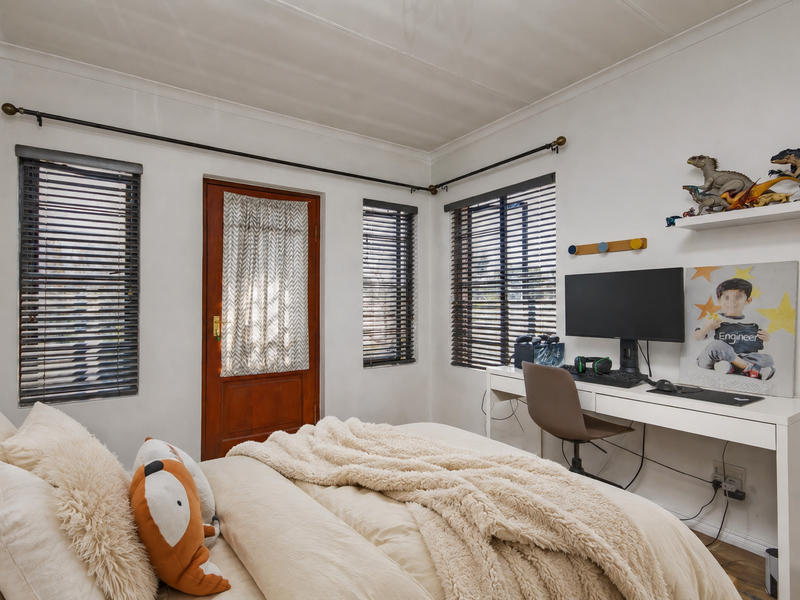 A well-lit bedroom featuring timber door access, multiple windows with venetian blinds, and integrated electrical points for a functional workspace.