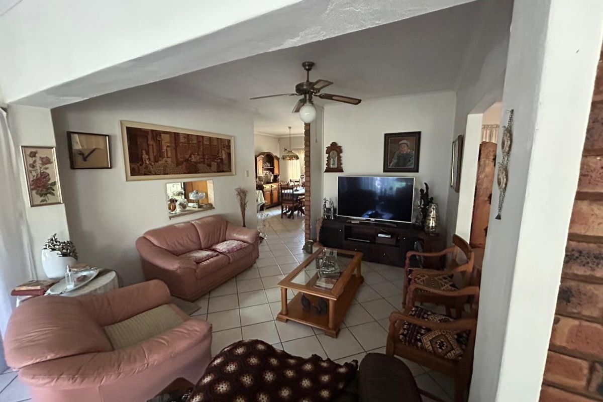 A comfortable living room featuring ceramic floor tiles, a ceiling fan, and an exposed brick pillar, with a pass-through window connecting to the dining area.