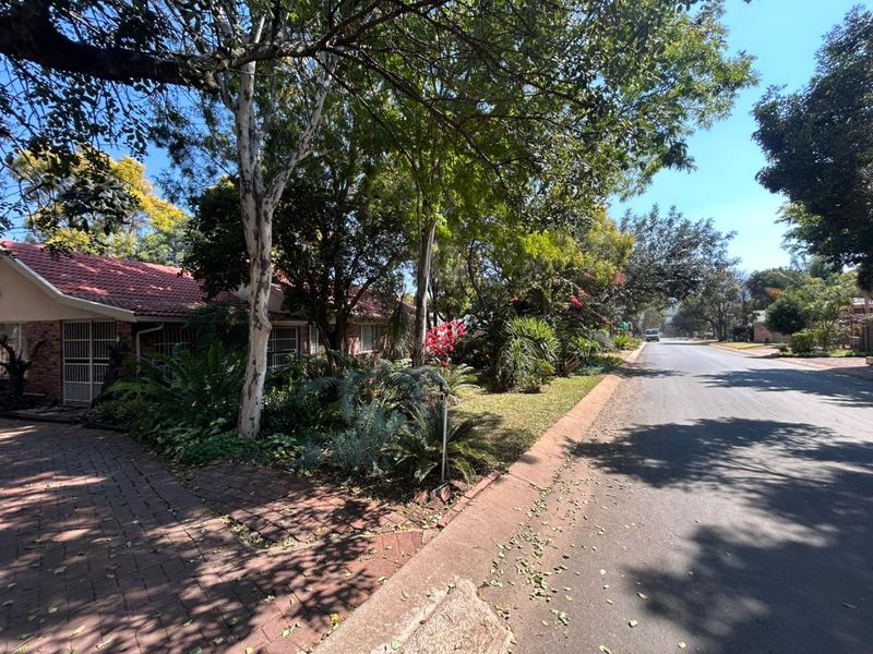 A street-level view of the property showing the paved driveway, brick facade, and mature garden landscaping along the road.
