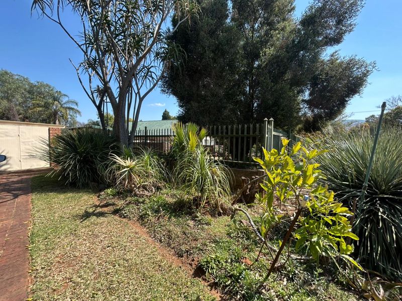 A landscaped garden area featuring established trees, shrubs, a brick driveway, and secure metal perimeter fencing.