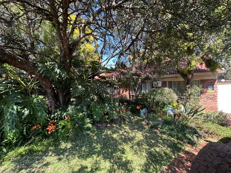 Front view of a brick house featuring a tiled roof, air conditioning unit, and a mature garden with established trees and paved driveway.