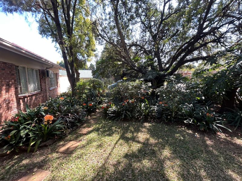 A lush, established garden area featuring mature trees and shrubs alongside the brick exterior of the house.