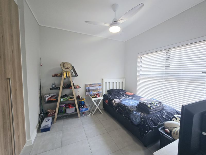 A well-maintained bedroom featuring neutral tiled flooring, a built-in wardrobe, a modern ceiling fan, and large windows fitted with venetian blinds.
