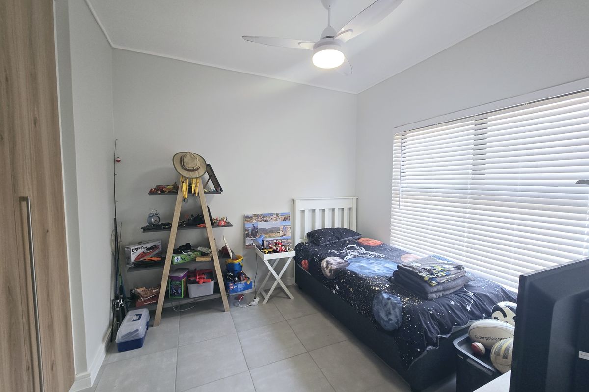 A well-maintained bedroom featuring neutral tiled flooring, a built-in wardrobe, a modern ceiling fan, and large windows fitted with venetian blinds.
