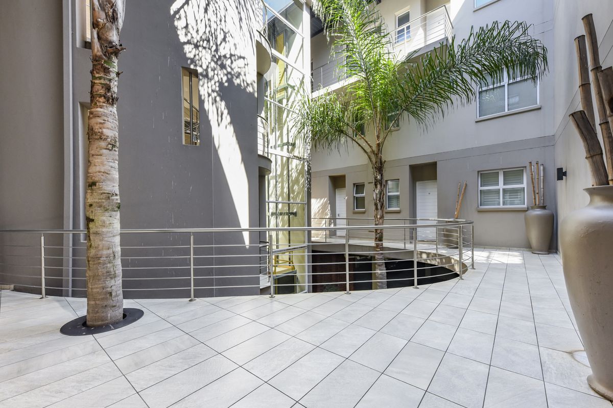 A modern apartment complex courtyard featuring tiled walkways, a glass elevator shaft, and stainless steel balustrades, providing access to multiple residential units.