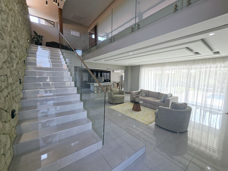Spacious open-plan living area featuring a striking stone feature wall, modern glass balustrade staircase, and high-quality tiled flooring with double volume ceilings.