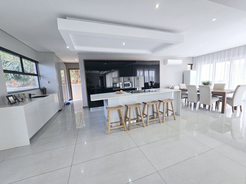 Modern open-plan kitchen and dining area featuring high-gloss cabinetry, a central island with seating, recessed ceiling lighting, and polished floor tiles.