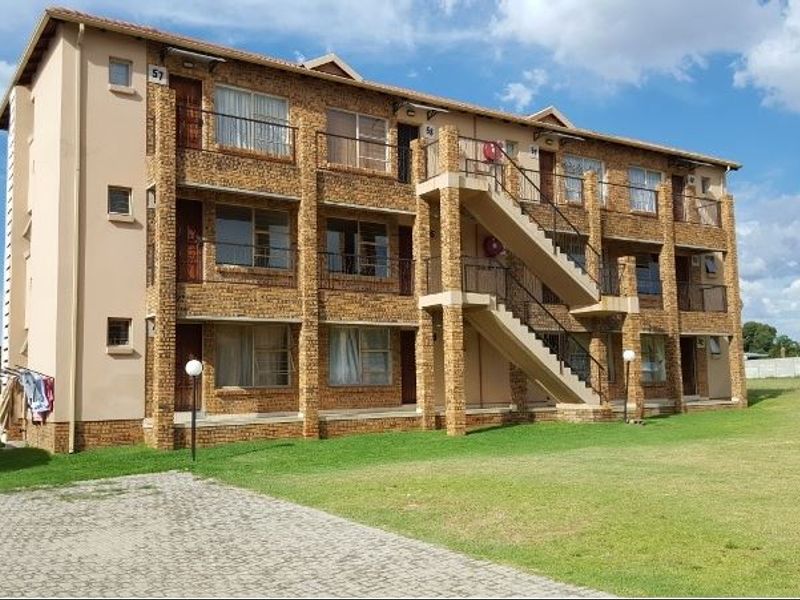 A multi-story apartment building featuring a face-brick facade, external staircases, and private balconies, set within a well-maintained communal garden area.