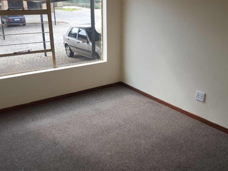 A clean, empty bedroom featuring neutral carpeted flooring, timber skirting boards, and a large window overlooking the parking area.