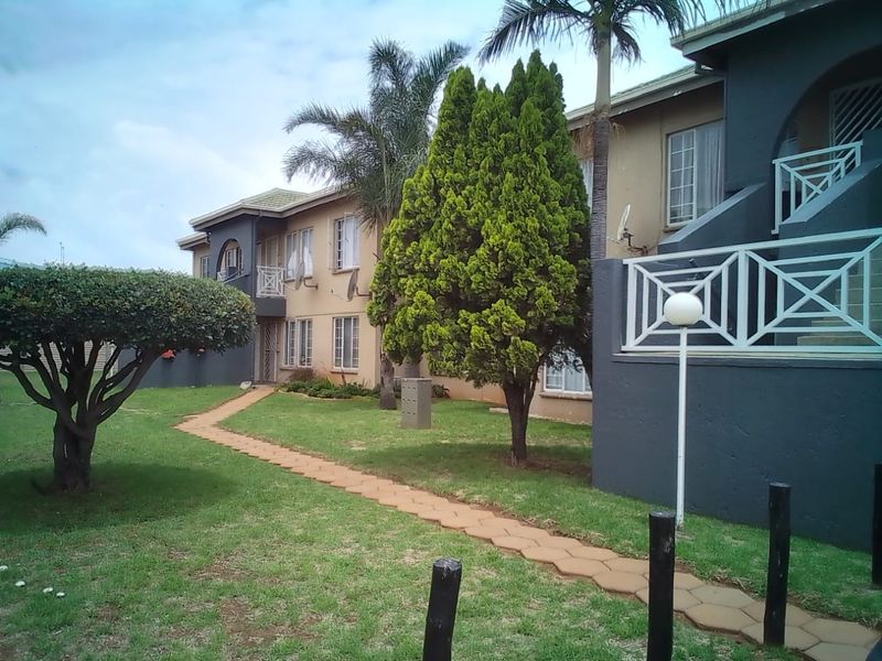 Exterior view of an apartment complex featuring a paved garden walkway, landscaped trees, and balcony access points.