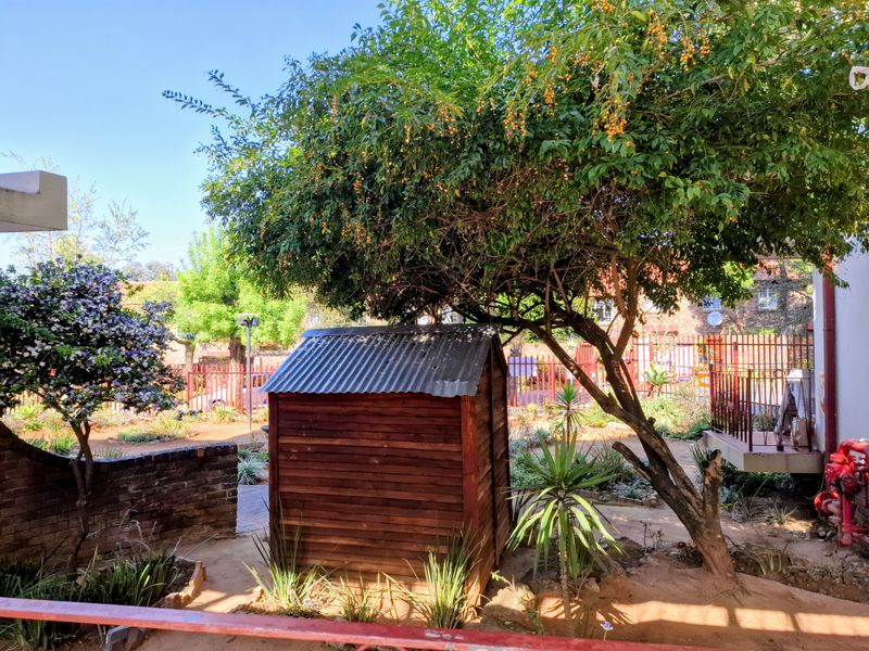 A communal garden area featuring a wooden storage shed, mature trees, brick retaining walls, and metal perimeter fencing.