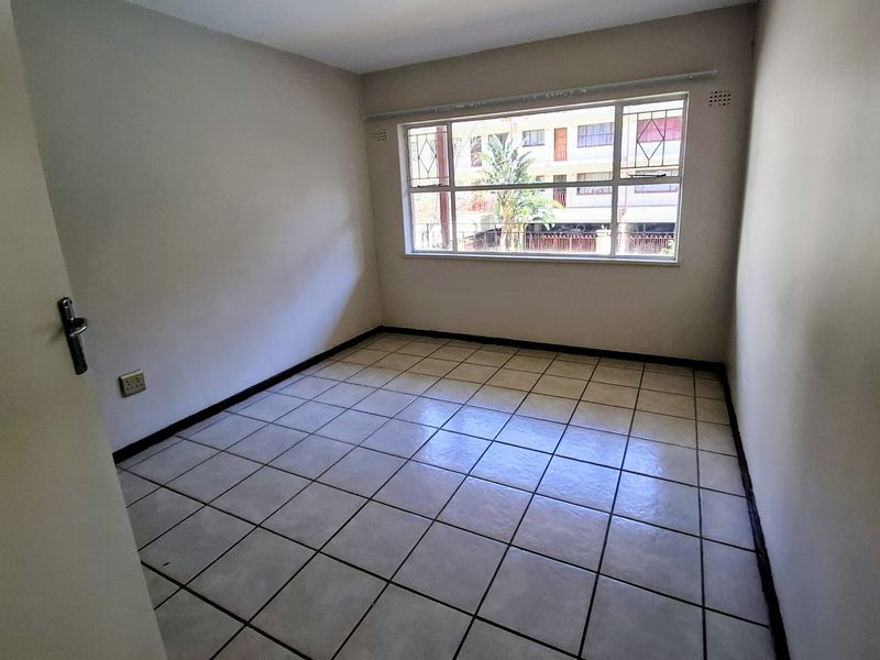 A bright, empty bedroom featuring neutral walls, tiled flooring, and a large window with security bars, offering a clean canvas for personalization.